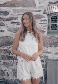 Hvit uten ermer topp med blonder og bølgede kanter, pairt med hvite shortser med intrikat broderi. Grå steinvegg i bakgrunnen.