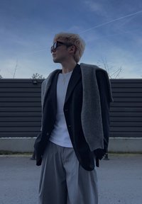 Personne aux cheveux blonds portant des lunettes de soleil, un blazer noir sur une chemise blanche, un pull gris drapé sur les épaules, et un pantalon gris ample en extérieur.