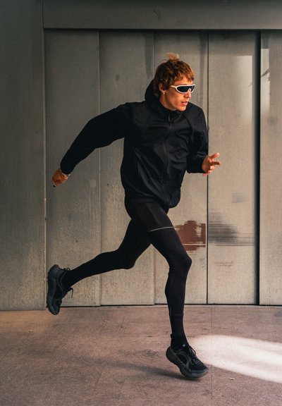 Corredor con ropa deportiva negra y gafas deportivas blancas trotando en interiores frente a una pared gris con paneles metálicos.