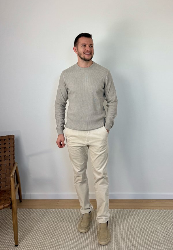 Man die een beige trui, crèmekleurige broek en beige schoenen draagt, staand op een licht tapijt naast een houten stoel tegen een effen witte muur.