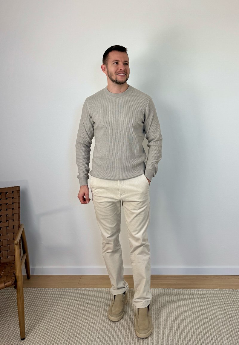 Homme portant un pull beige, un pantalon crème et des chaussures beiges, debout sur un tapis clair à côté d'une chaise en bois, devant un mur blanc uni.