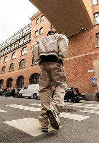 Homme traversant un passage pour piétons en ville près de bâtiments en briques, portant une veste beige avec un motif floral et un pantalon cargo beige.