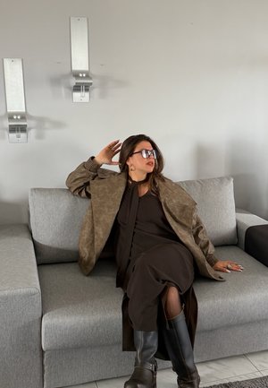 Mujer con gafas de sol, abrigo marrón, vestido negro y botas sentada en un sofá gris con la mano apoyada detrás de la cabeza y mirando hacia un lado.