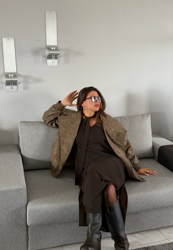Woman wearing sunglasses, brown coat, black dress, and boots sitting on grey sofa with hand resting behind head and looking to the side.