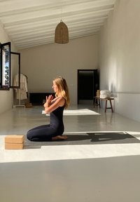 Praktyka jogi w jasnym pokoju, przedstawiająca osobę ubraną na czarno, klęczącą z dłońmi złączonymi przed sobą. Na podłodze leżą dwa bloki korkowe i mata do jogi.