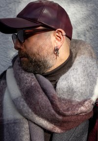 Viininpunainen nahkainen lakki, kilpikonnankuviolliset silmälasit, harmaa parta; päällä paksu, pehmeästi teksturoitu huivi, jossa sävyjä viininpunaisesta, harmaasta ja kermanvärisestä.