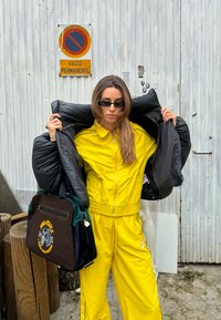 Κίτρινο φόρεμα γυμναστικής με φερμουάρ και ελαστική ζώνη στην μέση; υπερμεγέθη μαύρη γούνα ριγμένη. Καφέ τσάντα με πράσινες λεπτομέρειες και λογότυπο.