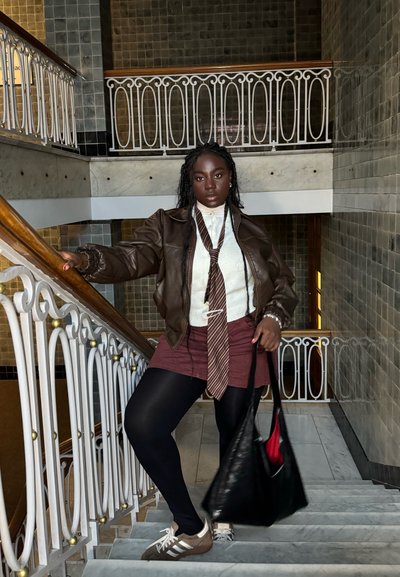 Chaqueta de cuero marrón sobre un suéter blanco, falda burdeos, medias negras y zapatillas marrones. Bolsa tote negra con interior rojo. Escalera decorativa.