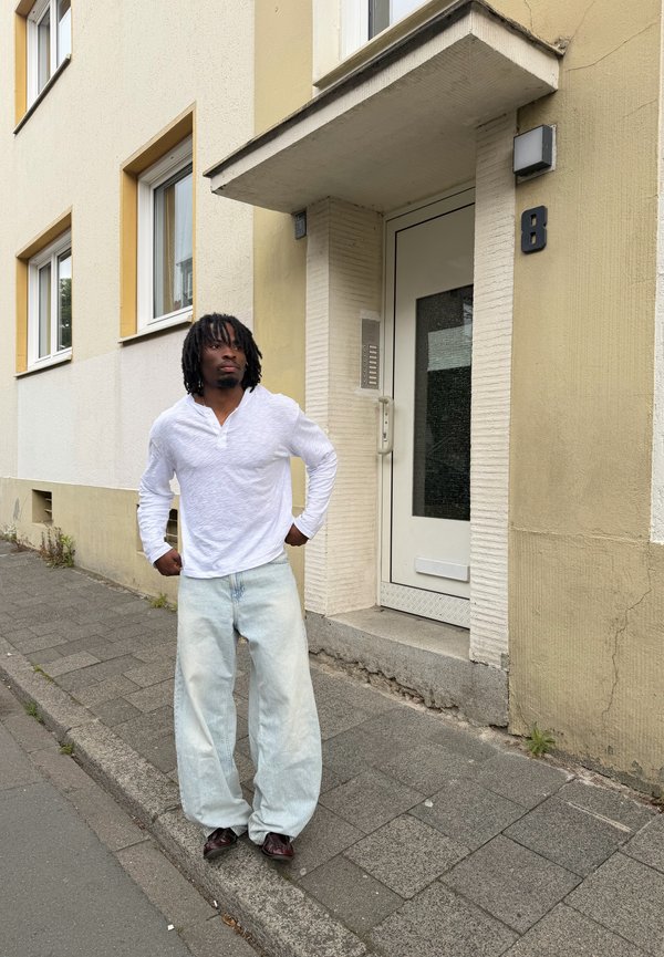 Homme avec des dreadlocks portant une chemise blanche à manches longues et un jean ample de couleur claire, debout sur le trottoir près d'une entrée de bâtiment beige avec le numéro de porte 8.