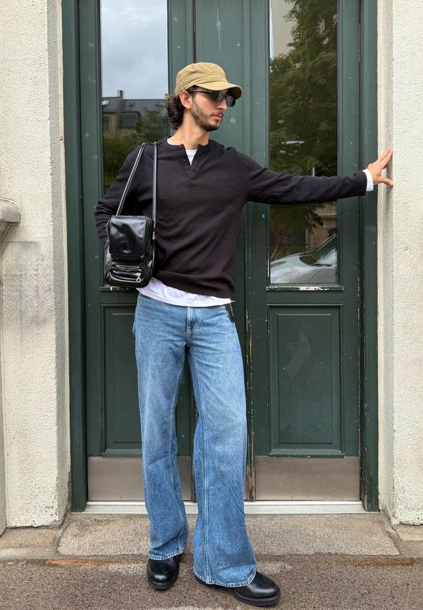 Uomo con berretto marrone chiaro, occhiali da sole, camicia nera, jeans blu e scarpe nere appoggiato a porte doppie verdi con una borsa nera sulla spalla.