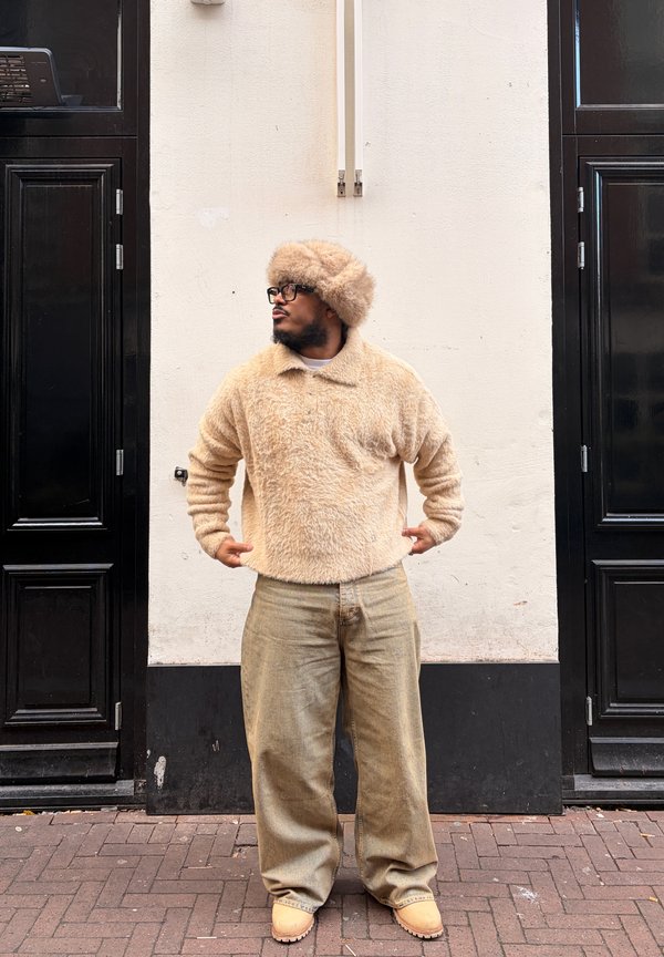 Homme portant un pull en laine beige, un chapeau assorti, un pantalon large beige et des chaussures beiges, debout entre deux portes noires sur un trottoir en briques.