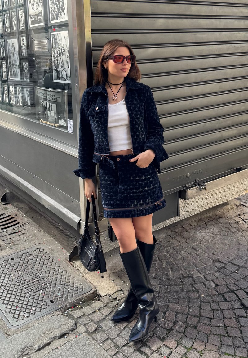 Femme en veste et jupe en velours noir, crop top blanc, bottes noires, lunettes de soleil et sac à main, debout sur une rue pavée près d'une boutique aux volets fermés.