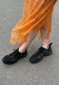 Svarta sportskor med texturerad meshöverdel och gummisula, tillsammans med en orange snörkjol. Synlig fot-tatuering på fotleden.