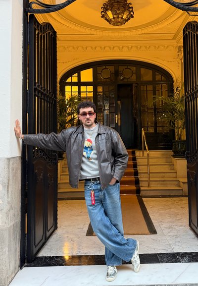 Hombre con gafas de sol, chaqueta de cuero y vaqueros, de pie de manera casual apoyado en una puerta de metal negro ornamentada, con suelo de mármol y escalera detrás.