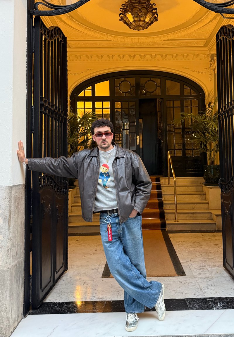 Mand iført solbriller, læderjakke og jeans står afslappet lænet op ad en smukt dekoreret sort metal døråbning med marmorgulv og trappe bagved.