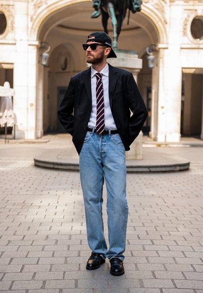 Hombre con gafas de sol, gorra al revés, chaqueta de traje, corbata rayada, vaqueros azul claro y zapatos negros, de pie con las manos en los bolsillos en una plaza.