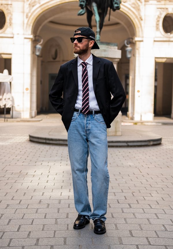 Man wearing sunglasses, backward cap, suit jacket, striped tie, light blue jeans, and black shoes standing with hands in pockets in a plaza.