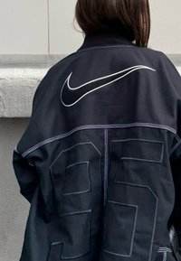 Personne portant une veste noire avec des coutures blanches, un grand logo Nike swoosh et le numéro 95 en relief dans le dos, debout contre un mur.