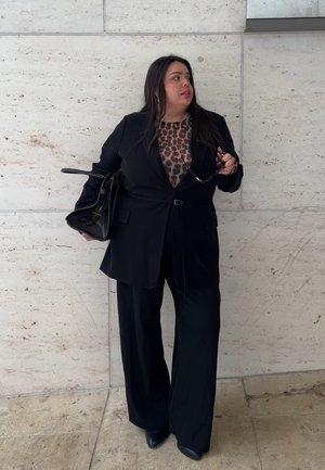 Mujer vestida con traje negro y blusa de estampado de leopardo sostiene una bolsa negra y gafas de sol, de pie contra una pared de piedra clara, mirando hacia un lado.