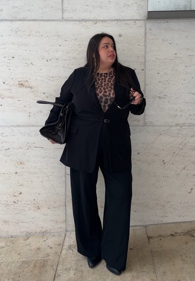 Mujer vestida con traje negro y blusa de estampado de leopardo sostiene una bolsa negra y gafas de sol, de pie contra una pared de piedra clara, mirando hacia un lado.