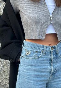 Γυναίκα που φοράει ανοιχτό γαλάζιο τζιν ψηλόμεσο, γκρι πουλόβερ με φερμουάρ που αφήνει ακάλυπτη την κοιλιά πάνω από λευκό τοπ, και μαύρο παλτό περασμένο σε ένα χέρι.