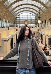 Karkea harmaa neulepaita, valkoisen ja sinisen raidallinen huivi sekä ruskea punottu laukku, taustalla koristeellinen museon sisustus.