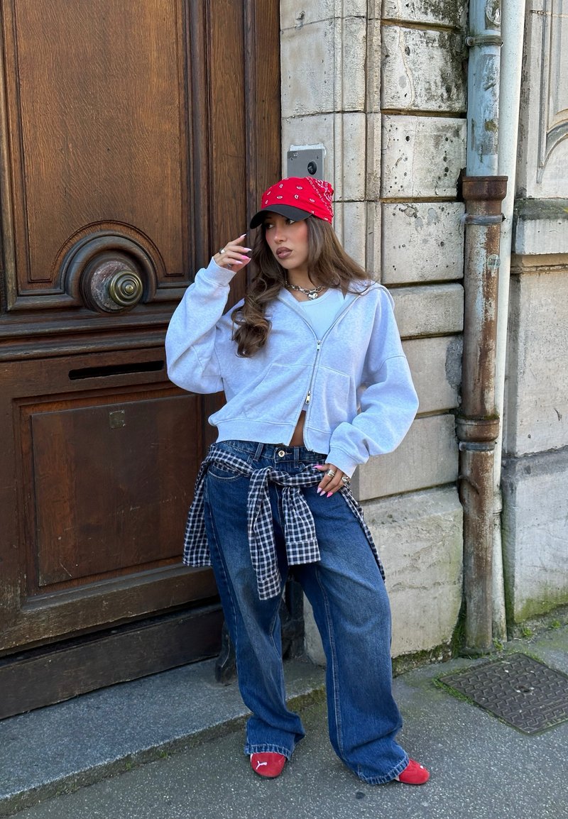 Giovane donna in abbigliamento casual con cappellino rosso, jeans oversize, felpa grigia con cappuccio e camicia a quadri annodata in vita, in piedi vicino a una porta di legno su una strada cittadina.