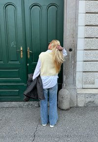 Kremowa, dziana kamizelka noszona na białej koszuli, połączona z dżinsami w kształcie dzwonów. Długie, proste blond włosy opadają na plecy. W tle ciemnozielone drzwi.