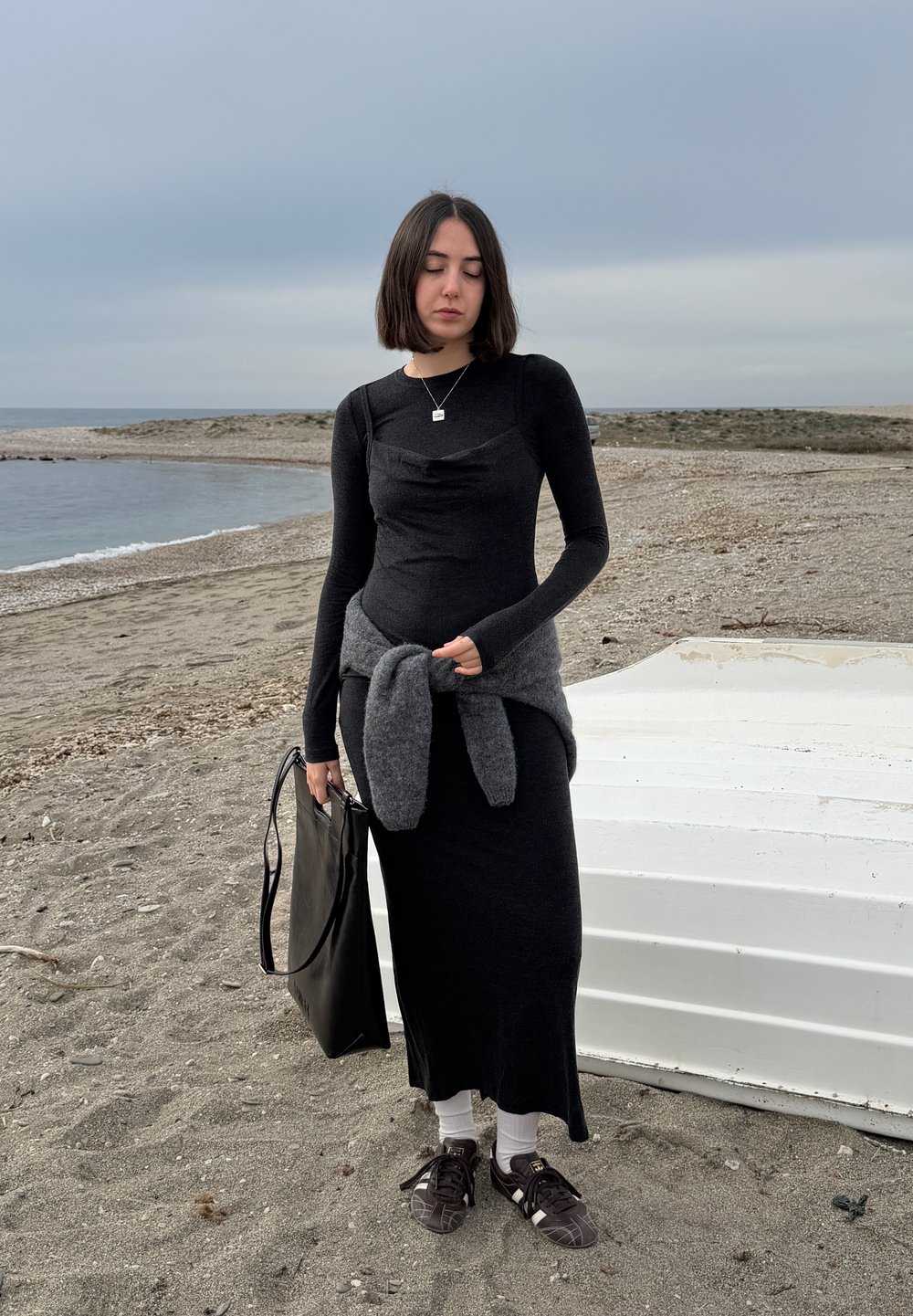 Jeune femme en robe noire et baskets se tient sur une plage de galets près d'un bateau blanc retourné sous un ciel nuageux.