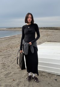Jeune femme en robe noire et baskets se tient sur une plage de galets près d'un bateau blanc retourné sous un ciel nuageux.