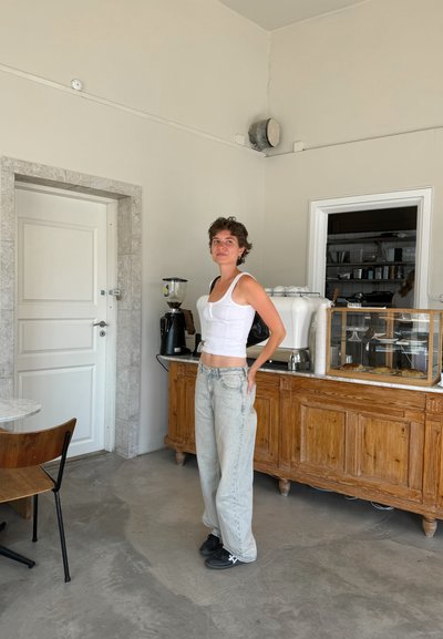 Top sin mangas blanco ajustado; jeans anchos azul claro; zapatillas negras. Mostrador de madera con una vitrina en un interior de colores claros.