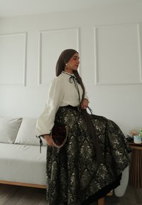 Blouse blanche à manches bouffantes avec détail de nœud noir, associée à une jupe longue noire à motifs dorés. Accessoirisée d'un sac à main sombre.
