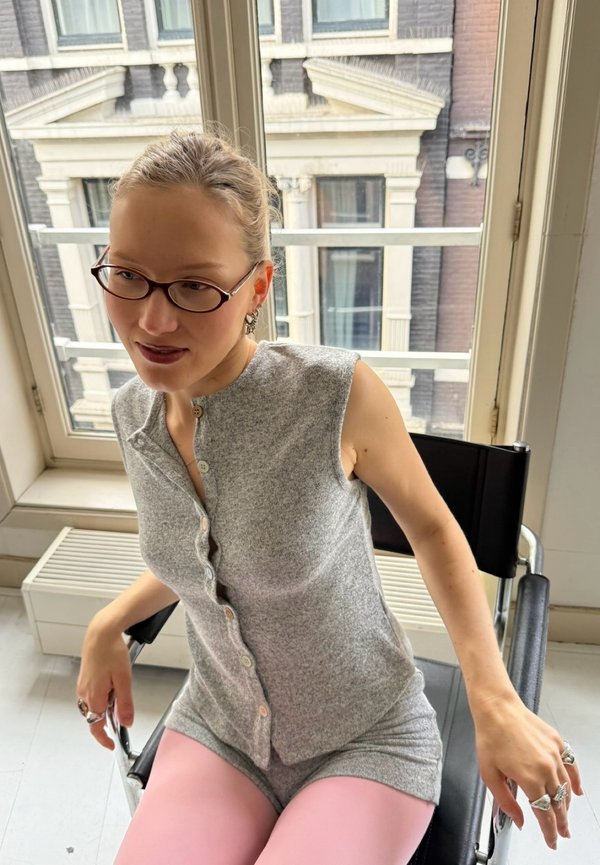 Frau mit Brille und Creolen, sitzt auf einem schwarzen Stuhl nahe einem Fenster, trägt ein graues ärmelloses Oberteil und rosa Leggings.