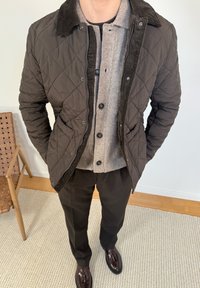 Homme portant une veste matelassée marron par-dessus un cardigan beige boutonné, un pantalon foncé, et des mocassins marron à pampilles, debout à l'intérieur près d'une chaise en bois.