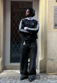 Casaco de treino preto da Adidas com riscas brancas, jeans largos pretos e sapatos pretos. Aposicionado em frente a uma porta com uma janela gradeada.