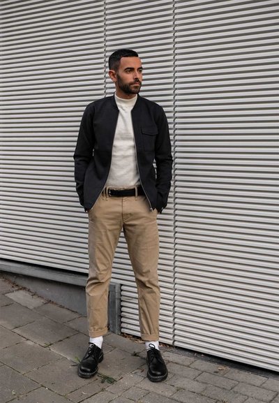 Chaqueta negra con cremallera sobre un suéter crema, pantalones caqui y zapatos negros. Calcetines blancos visibles. De pie frente a un fondo gris texturizado.