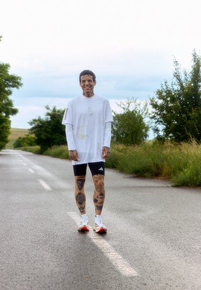 Atleta con una camiseta blanca de corte holgado con gráficos, pantalones cortos negros de Adidas y zapatillas blancas con detalles rojos, de pie sobre una carretera asfaltada.