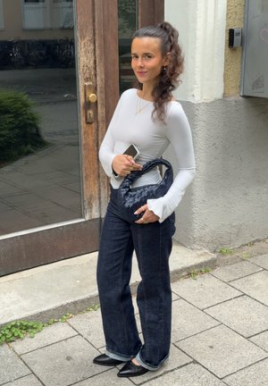 Top blanco de manga larga con mangas acampanadas, pantalones vaqueros oscuros de pierna ancha y un bolso negro trenzado. Fondo de hormigón con una puerta de madera.