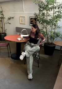 Ένα καθιστό άτομο φοράει ένα εφαρμοστό μπλουζάκι με μακριά μανίκια σε μαύρο και καφέ χρώμα, φαρδιά ανοιχτό πράσινα παντελόνια και λευκά αθλητικά παπούτσια, κρατώντας ένα φλιτζάνι σε ένα ξύλινο τραπέζι.