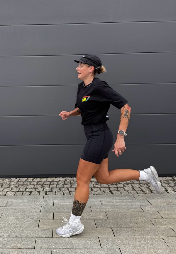 Frau läuft draußen und trägt schwarze Sportkleidung, weiße Sneakers, eine Kappe und sichtbare Tattoos an Bein und Arm.