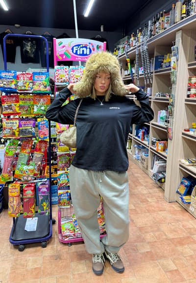 Camisa negra de manga larga con logo, pantalones de chándal grises, sudadera de pelaje sintético, bolso beige, de pie en una tienda de conveniencia con estanterías de caramelos detrás.