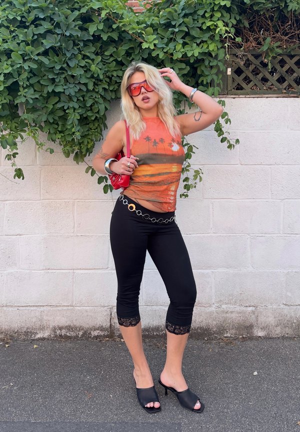 Frau mit blondem Haar, die rot getönte Sonnenbrillen, ein oranges ärmelloses Oberteil mit Palmen, schwarze gekürzte Hosen und schwarze Absätze trägt und an einer Wand steht.