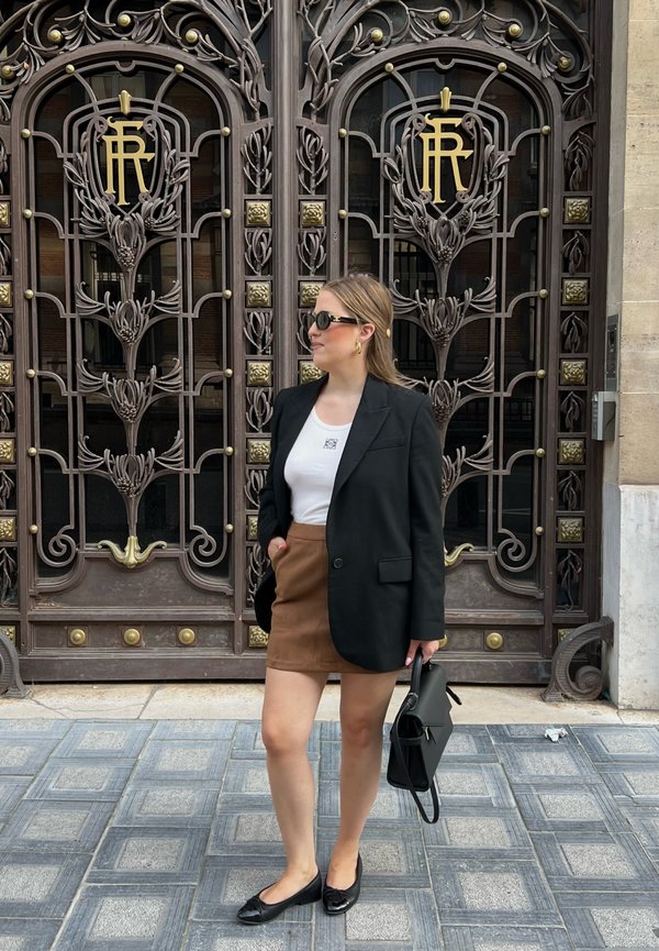 Blazer nero su una camicia bianca, gonna mini marrone e ballerine nere. Accenti dorati su un cancello in ferro battuto sullo sfondo. Piastrelle quadrate sotto i piedi.