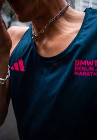 Futó sötétkék atlétatrikót visel, amelyen a "BMW Berlin Marathon" logó látható, valamint egy ezüst nyakláncot csillag alakú medállal.