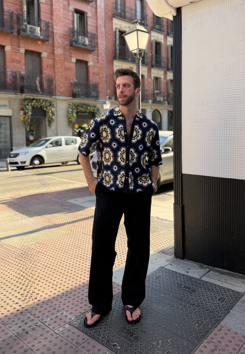 Homme portant une chemise à manches courtes à motifs, un pantalon noir et des sandales, debout sur un trottoir en ville près d'un coin de bâtiment.