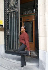 Maglione a maglia bordeaux con maniche lunghe, pantaloni neri a campana e tacchi alti neri, in piedi su gradini di pietra accanto a una porta in metallo decorata.