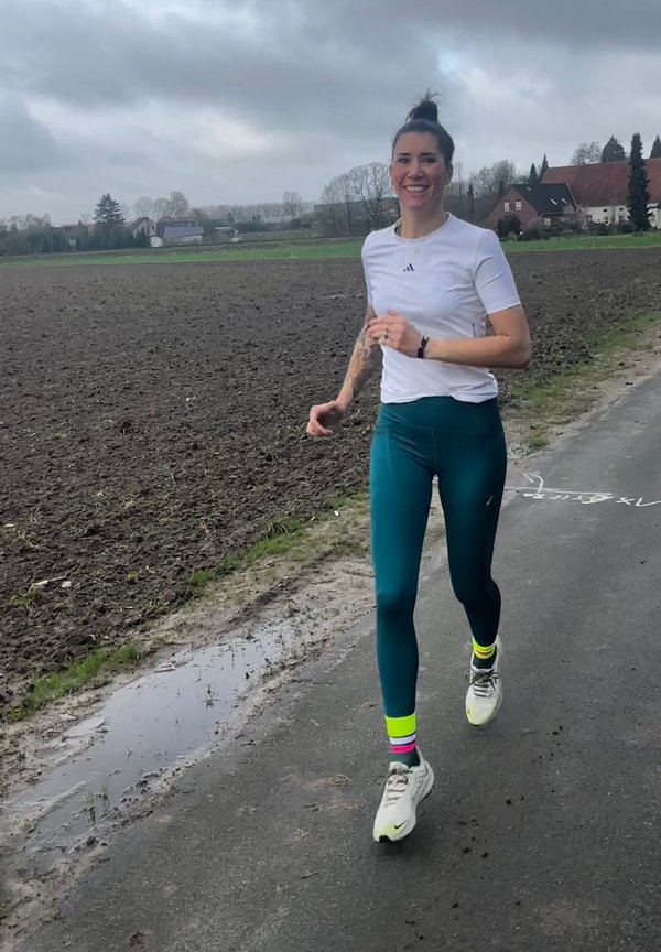 Atletinja v beli majici s kratkimi rokavi in teal legicah z rožnatimi in rumenimi nogavicami do gležnjev, teče po poti ob blatnem polju.