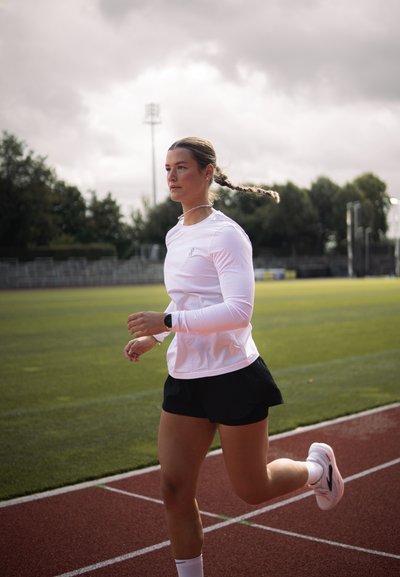 Camiseta atlética blanca de manga larga y ligera, pantalones cortos negros para correr, reloj plateado; corredor en una pista con fondo de césped verde y cielo nublado.