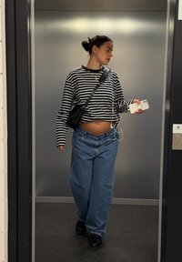 Fekete-fehér csíkos hosszú ujjú ing, bő kék farmernadrág és fekete tornacipő. Egy fehér telefont tart fülhallgatóval a kezében, egy liftben.