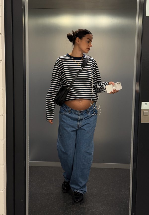 Fekete-fehér csíkos hosszú ujjú ing, bő kék farmernadrág és fekete tornacipő. Egy fehér telefont tart fülhallgatóval a kezében, egy liftben.